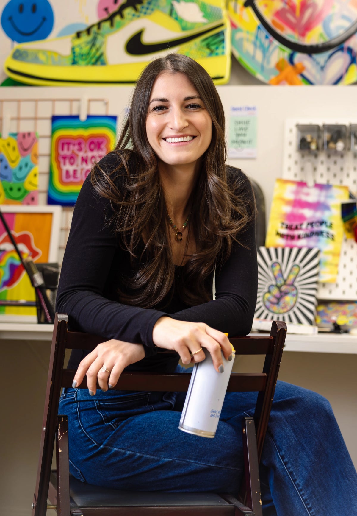 Woman sitting on a chair holding a can of spray paint in an art classroom with colorful artwork on the walls.