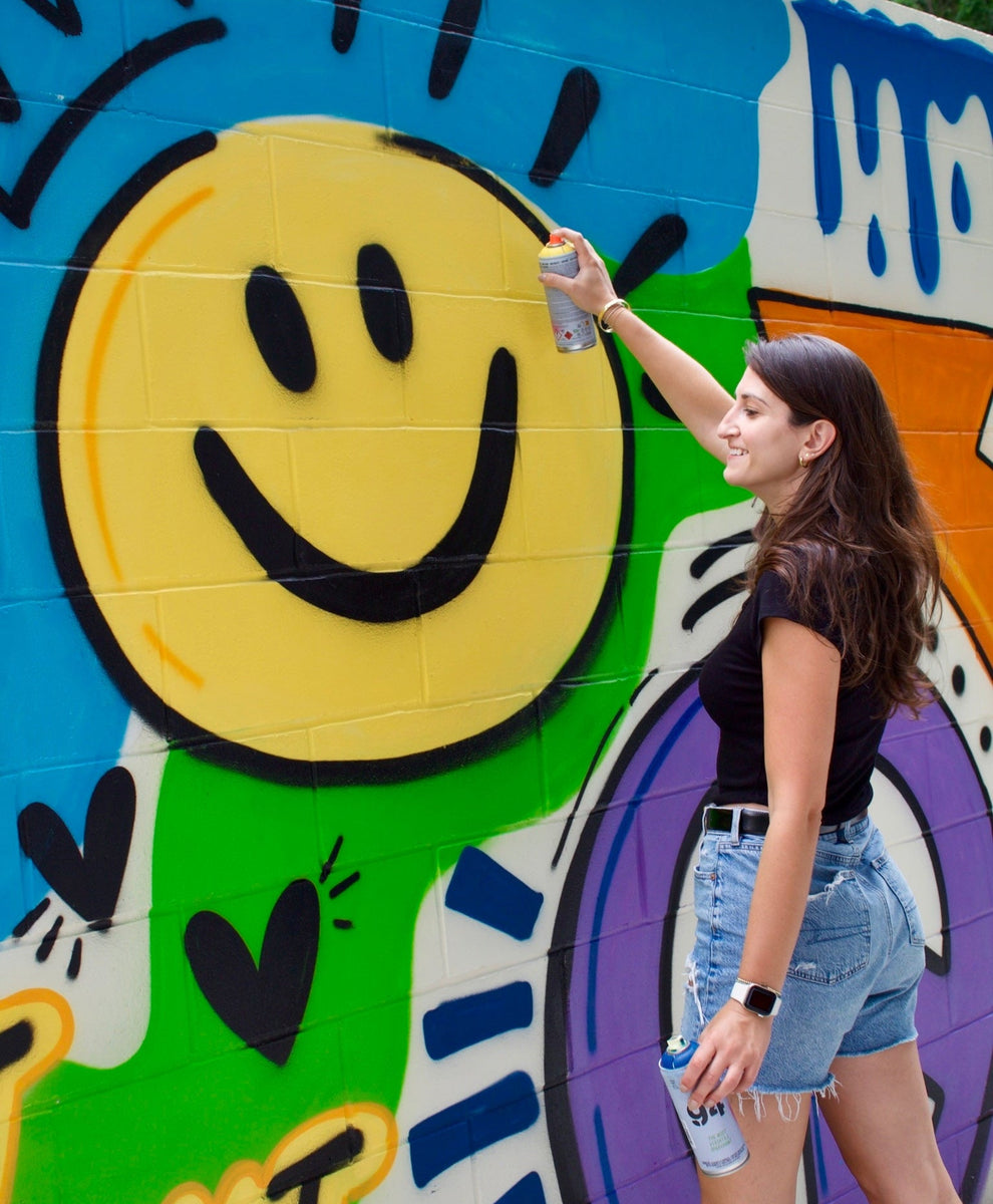 Person spray painting a colorful mural with a smiley face on a wall.