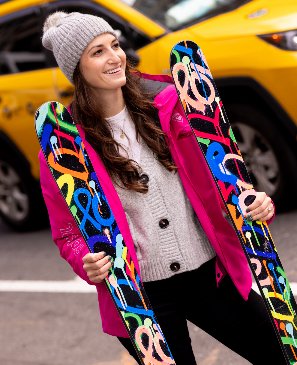 Woman holding colorful skis with a pink coat and gray beanie in an urban setting.